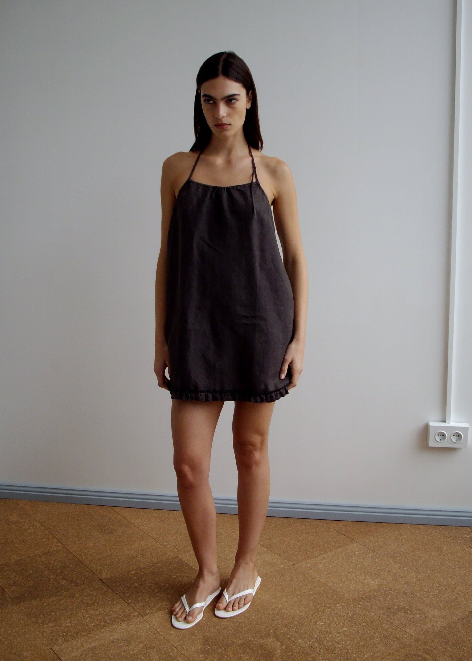 A woman with straight dark hair stands indoors against a plain white wall, wearing the Flounce Halter Dress - Bark and white flip-flops. She has a serious expression and her arms rest relaxed by her sides.