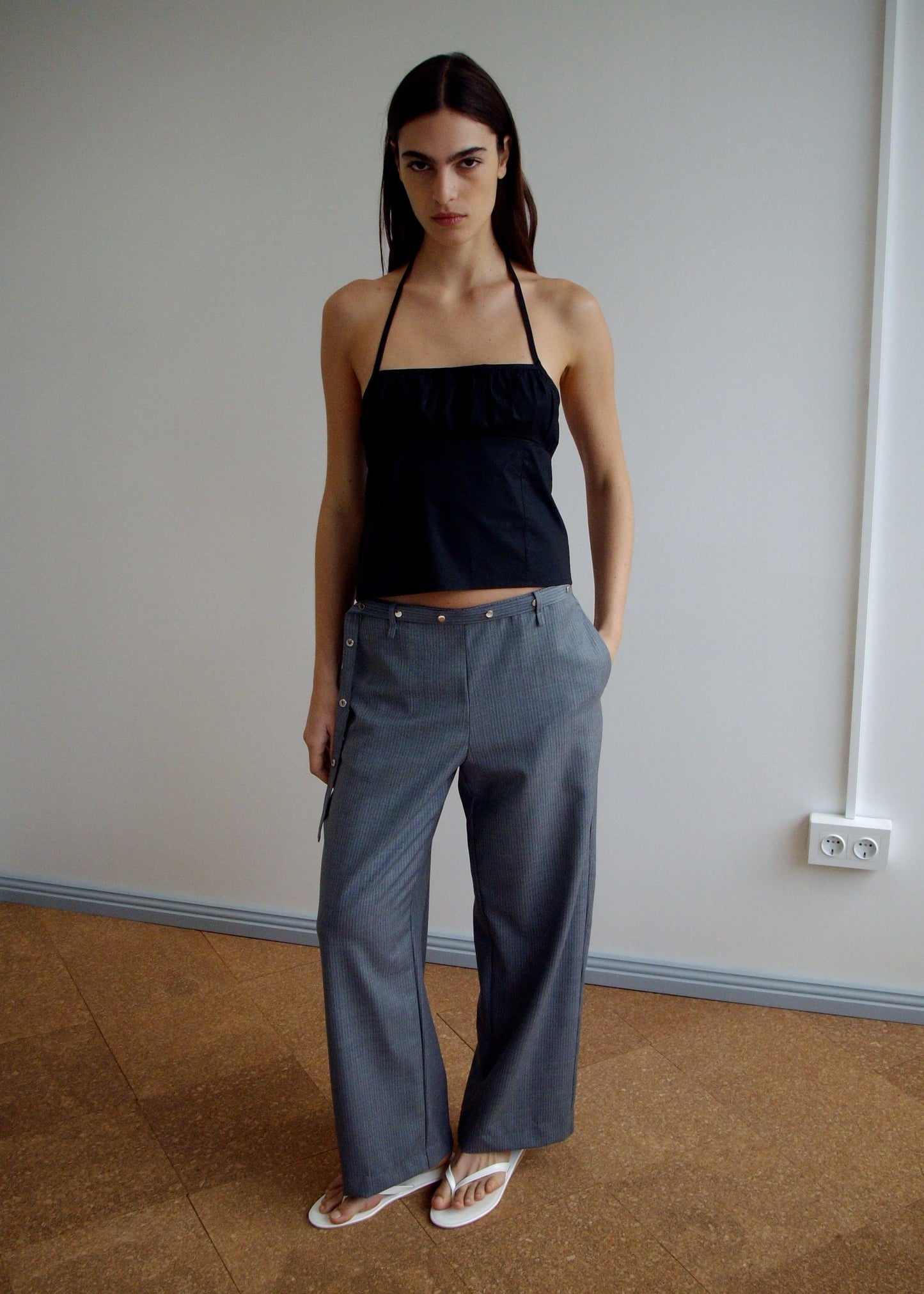 A person stands indoors against a plain wall wearing a Deiji Studios Strap Top - Black, gray wide-leg pants with zippers, and white flip-flops, hands in pockets and a neutral expression.