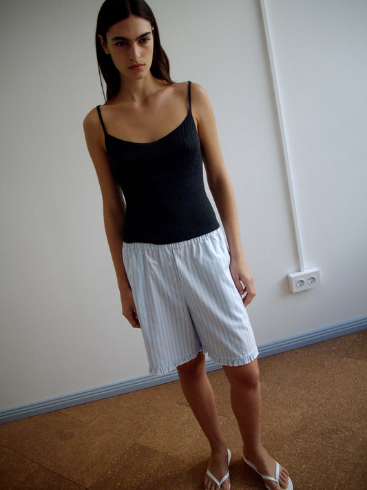 A person stands indoors wearing a black tank top, Flounce Short - Mineral Stripe, and white sandals, with a white wall, electrical outlet, and brown flooring in the background.