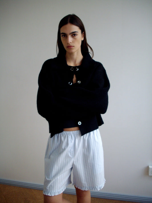 Indoors against a plain wall, a person with long dark hair and a neutral expression wears the oversized Stud Cardi Knit - Black, made from recycled materials, paired with loose, light-colored striped shorts, arms crossed.