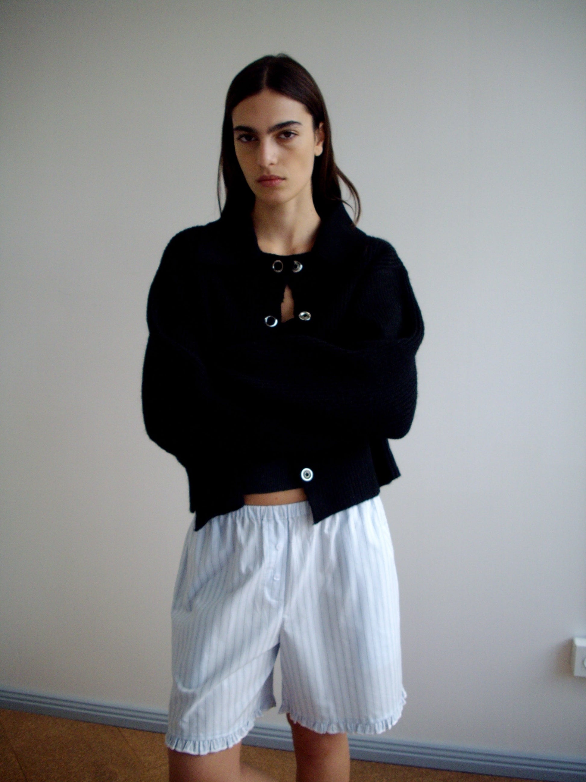 A person with long dark hair stands indoors against a plain wall, wearing a black buttoned sweater and the Flounce Short - Mineral Stripe, arms crossed and a neutral expression.