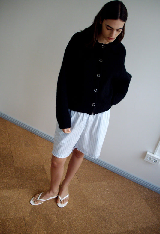 Indoors, a person stands against a plain wall and brown floor wearing the Stud Cardi Knit - Black, styled oversized with loose white striped shorts and white sandals, looking down with hands behind their back.