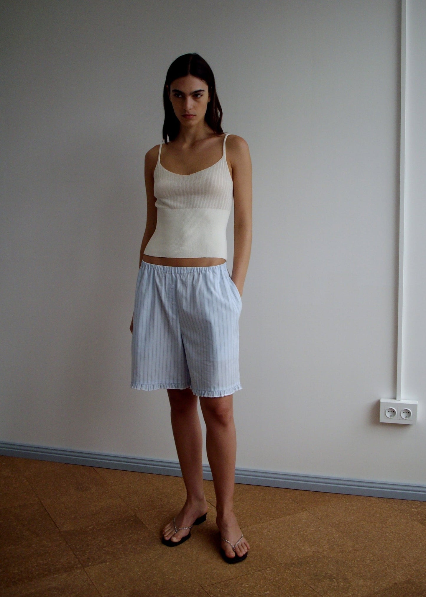 A woman with long dark hair stands indoors against a plain wall, wearing the Deiji Studios Ribbed Tank - Off White, loose light blue striped lyocell shorts, and brown sandals. She has a neutral expression.