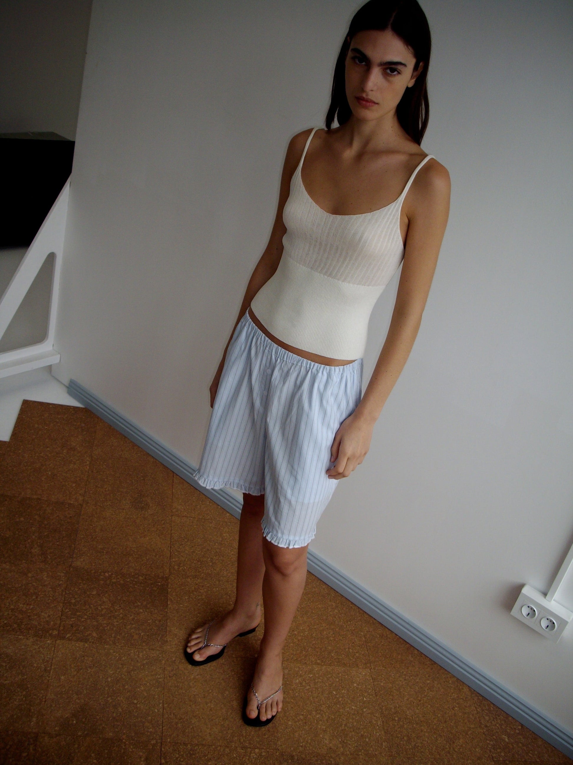 A woman with long dark hair stands indoors, wearing the Ribbed Tank - Off White by Deiji Studios and light blue striped shorts. She looks at the camera with a neutral expression in a minimally decorated, softly lit setting.