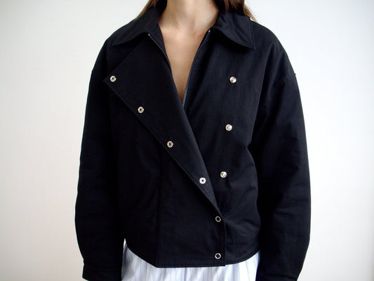 A person models the Lapel Jacket - Black with asymmetric lapels and shiny silver buttons, partially unzipped over a white outfit against a plain light background, highlighting its V neckline.