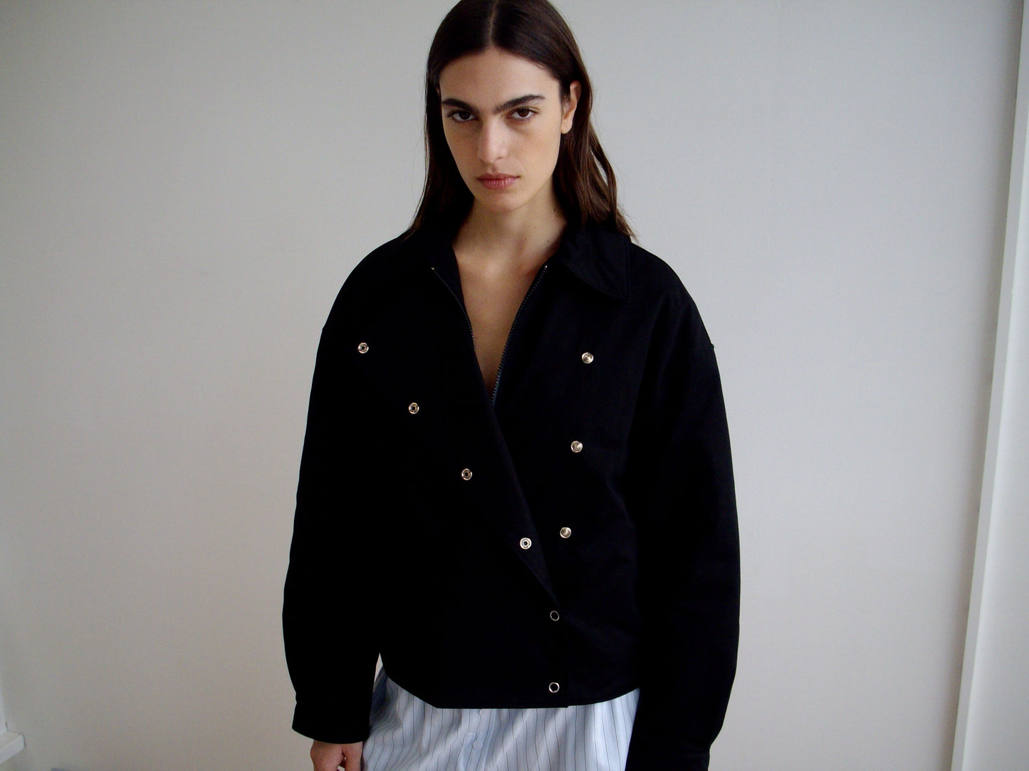 A person with long dark hair stands indoors against a plain white wall, wearing the Lapel Jacket - Black from Deiji Studios and light pants, gazing seriously at the camera.