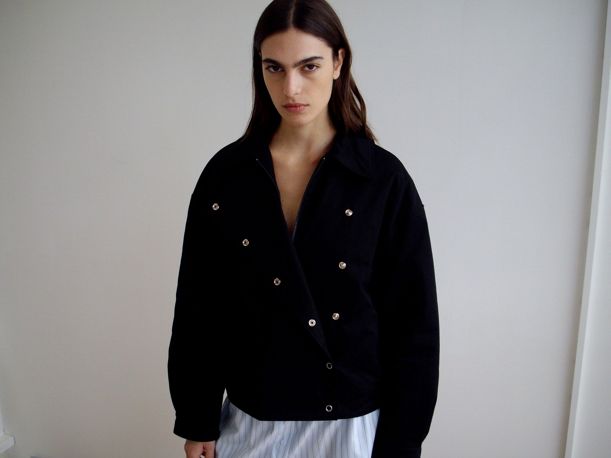 A person with long dark hair stands indoors against a plain white wall, wearing the Lapel Jacket - Black from Deiji Studios and light pants, gazing seriously at the camera.