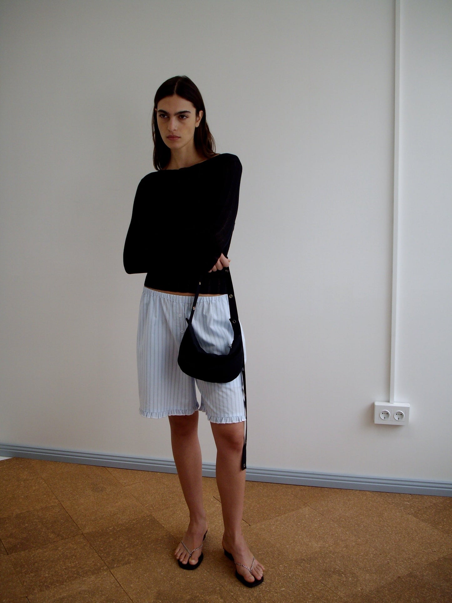 A woman with straight dark hair and a neutral expression stands indoors against a plain wall, wearing a black long-sleeve top, loose white striped shorts from Deiji Studios, black sandals, and holding the Snap Belt Bag - Black.