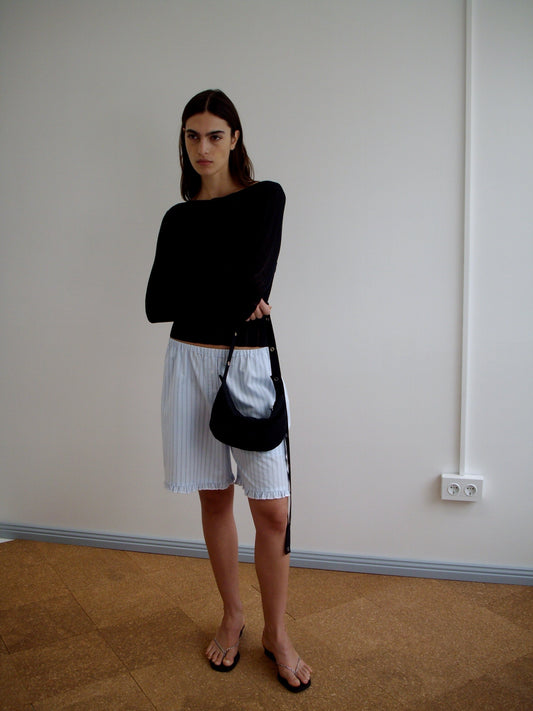 A woman with straight dark hair and a neutral expression stands indoors against a plain wall, wearing a black long-sleeve top, loose white striped shorts from Deiji Studios, black sandals, and holding the Snap Belt Bag - Black.