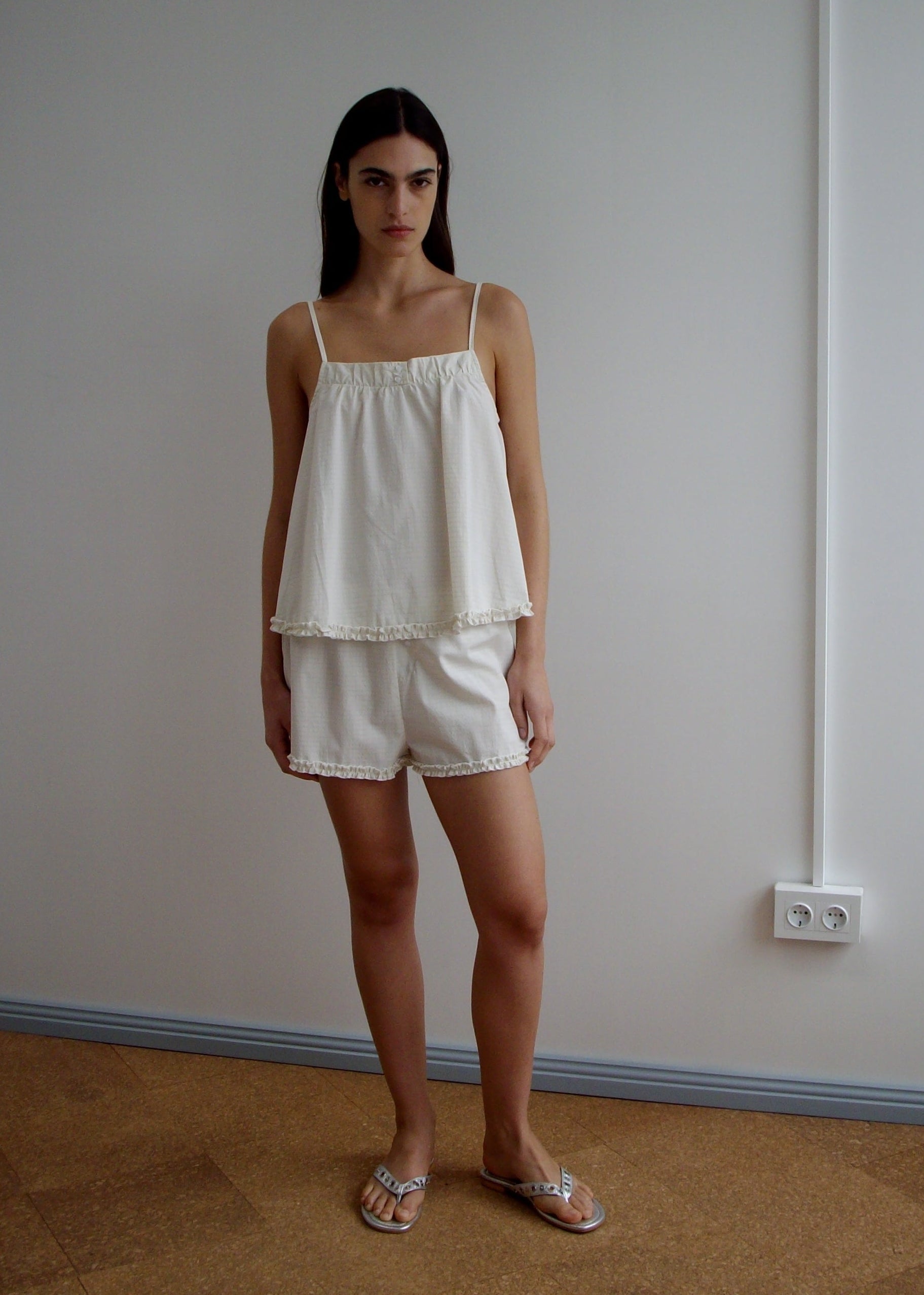 A woman with long dark hair and a neutral expression stands indoors against a plain white wall, wearing the Yoke Set - Bone Check, a sleeveless, loose-fitting organic cotton poplin babydoll top with matching shorts and sandals.