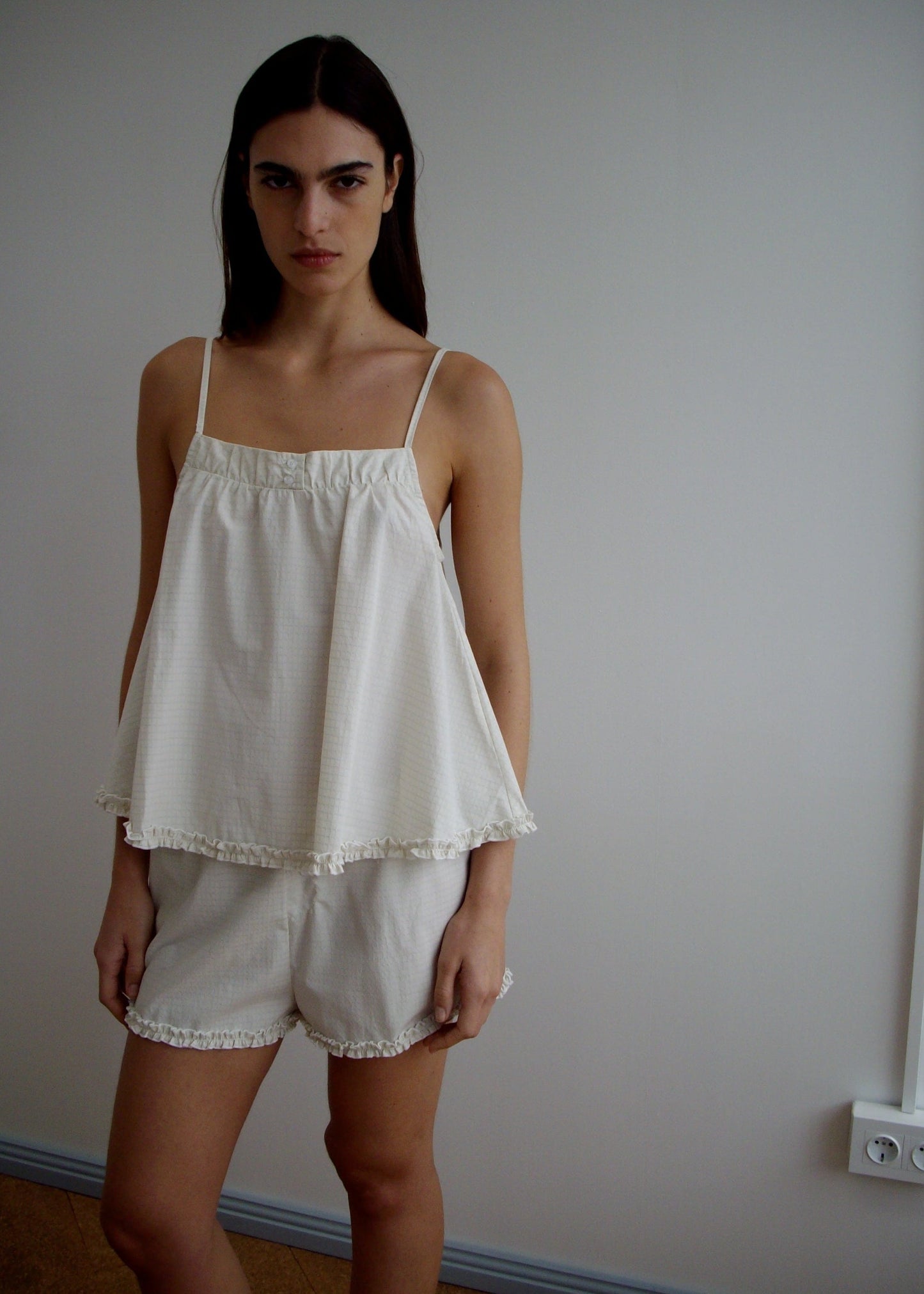 A woman with long dark hair stands indoors wearing the Yoke Set - Bone Check, a sleeveless, loose-fitting babydoll top and matching shorts in organic cotton poplin with ruffled edges, set against a plain light-colored wall.