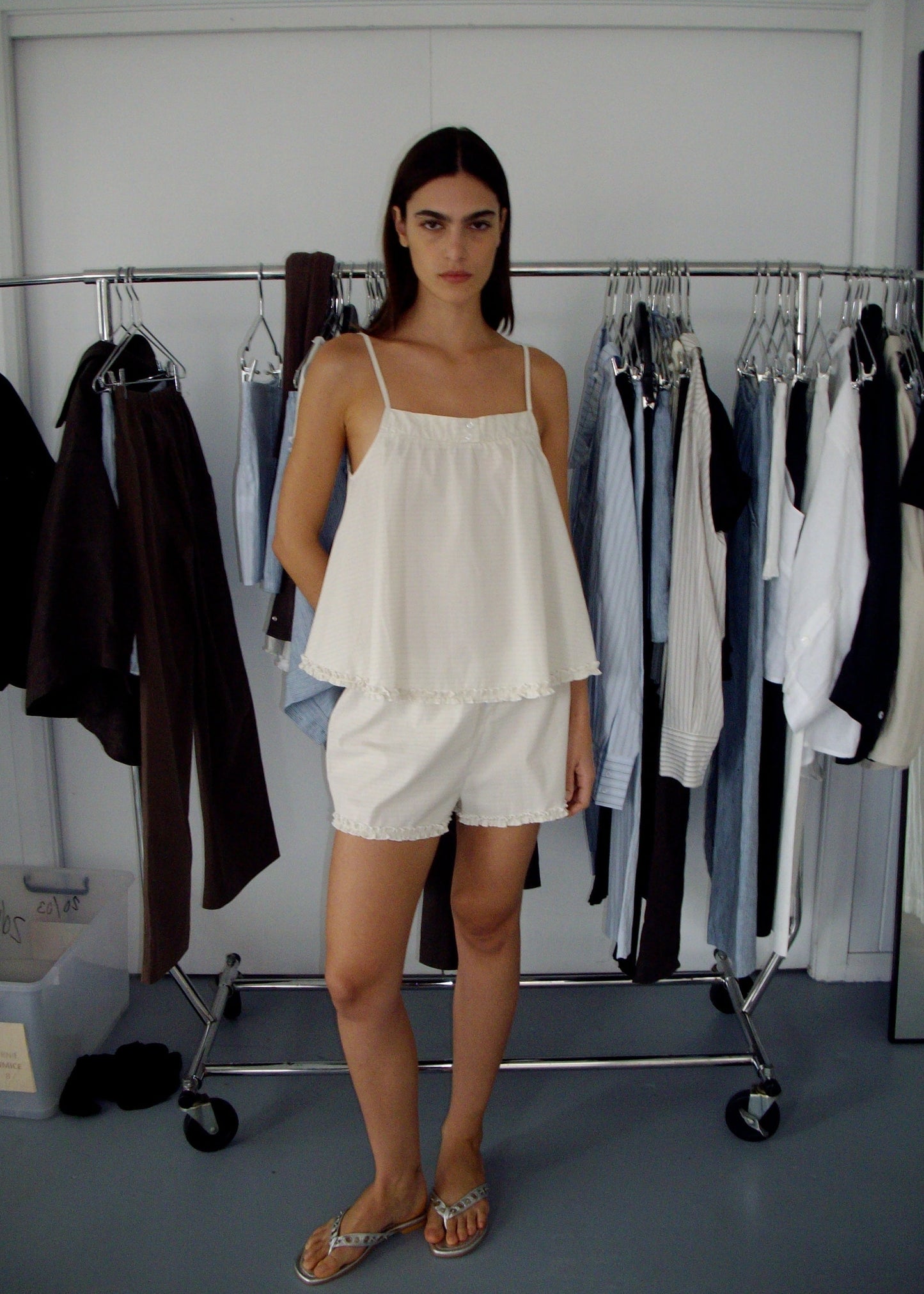 A woman stands indoors in front of two clothing racks, wearing the Yoke Set - Bone Check, featuring a loose organic cotton poplin babydoll top and matching shorts. The racks behind her display garments in dark and light neutral tones.
