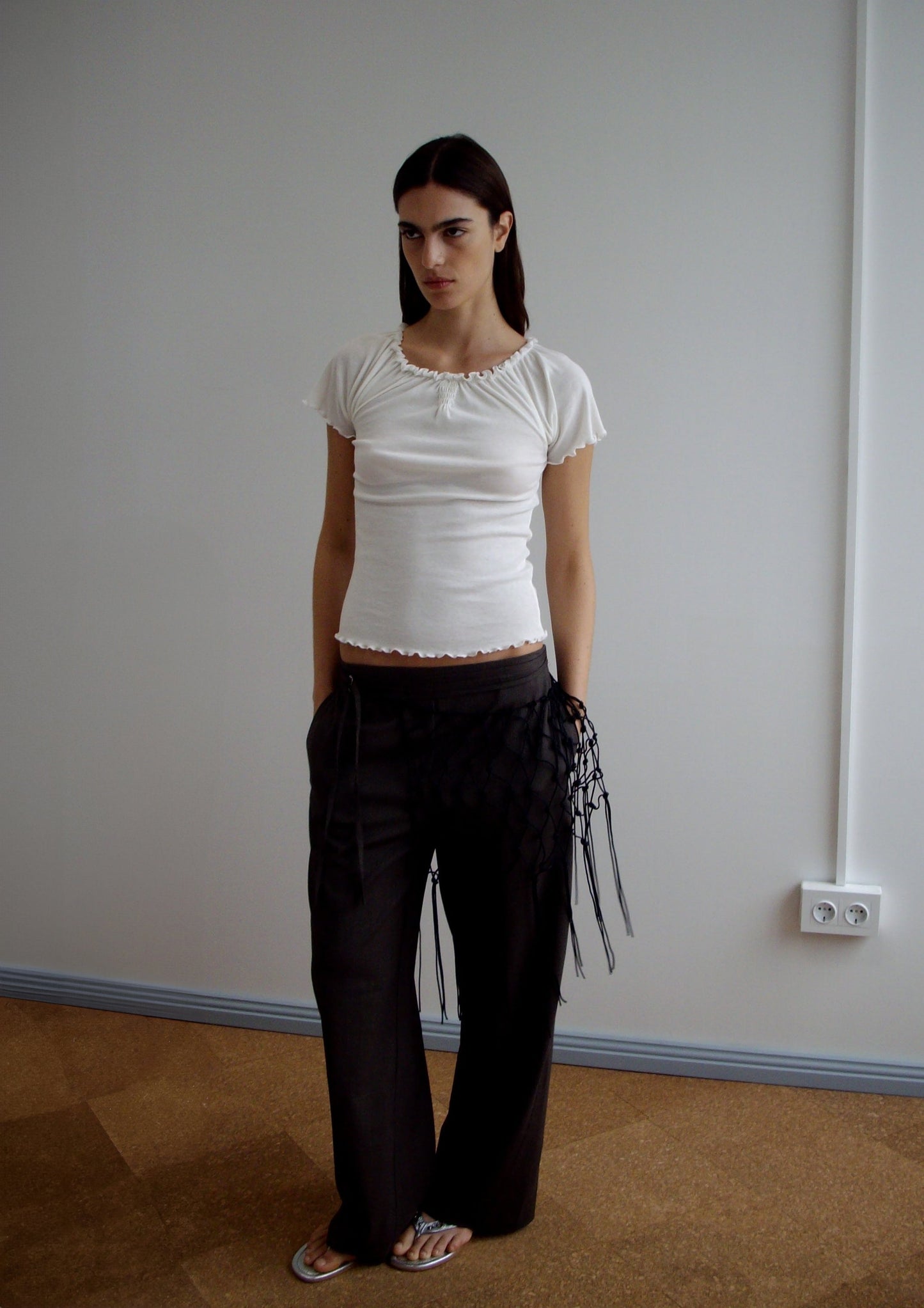 A woman stands indoors against a plain wall, wearing the Flounce Tee - Cream by Deiji Studios, loose dark pants with black fringe details, and white flip-flops. She has long dark hair and a neutral expression.