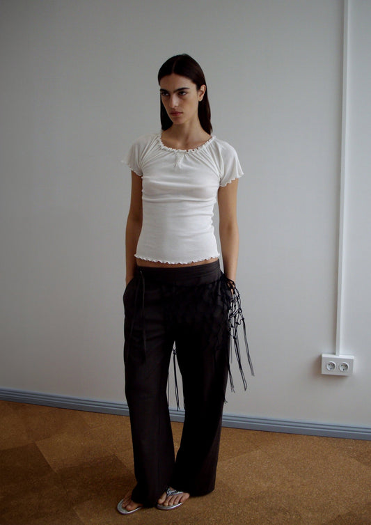 A woman stands indoors against a plain wall, wearing the Flounce Tee - Cream by Deiji Studios, loose dark pants with black fringe details, and white flip-flops. She has long dark hair and a neutral expression.