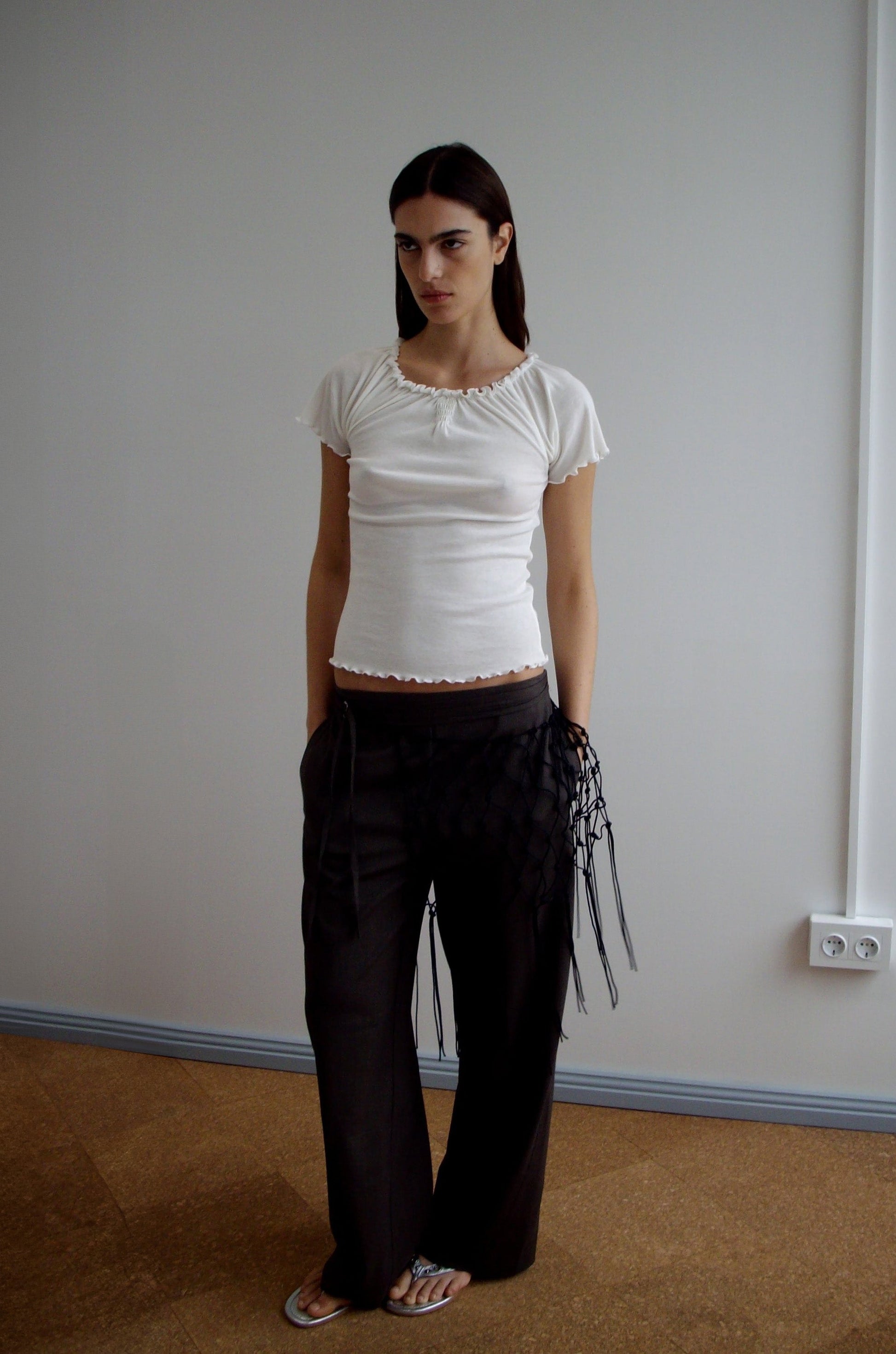 A woman stands indoors against a plain wall, wearing a fitted white short-sleeve top, loose dark pants accessorized with the Macrame Belt - Black, and sandals. Her hands are in her pockets and she maintains a neutral expression.