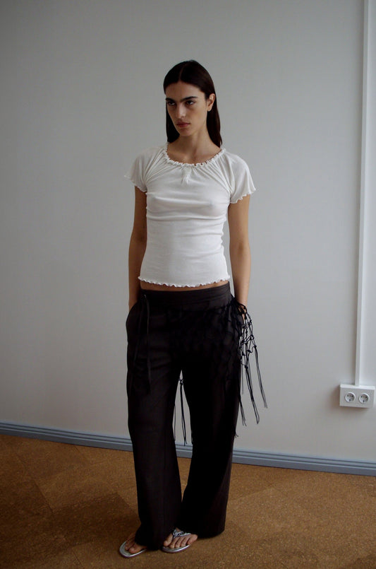 A woman stands indoors against a plain wall, wearing a fitted white short-sleeve top, loose dark pants accessorized with the Macrame Belt - Black, and sandals. Her hands are in her pockets and she maintains a neutral expression.