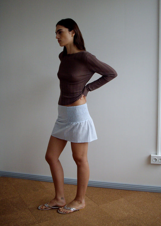 A woman with long dark hair stands sideways indoors, wearing a brown top, sandals, and the Deiji Studios Shirring Skirt - Mineral Stripe. She rests her hands on her lower back by a plain wall with an electrical outlet.
