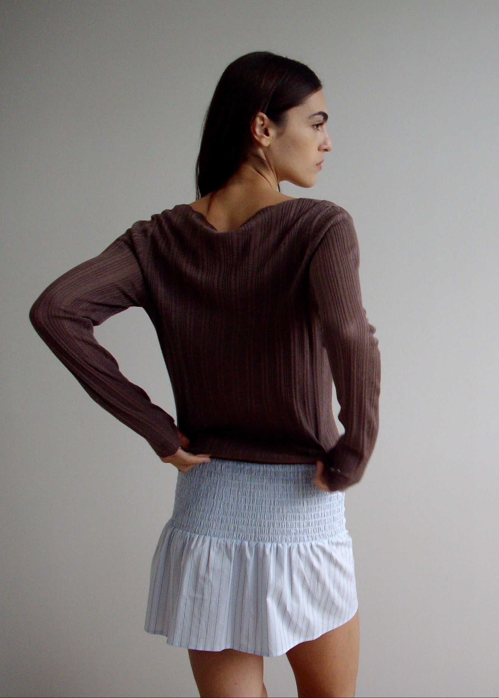 A woman faces sideways with her head turned, wearing a brown long-sleeve top and the Shirring Skirt—a light blue smocked mini skirt with ruffles—against a plain, light-colored background.