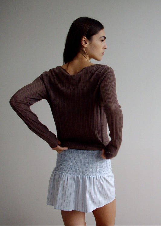 A woman faces sideways with her head turned, wearing a brown long-sleeve top and the Shirring Skirt—a light blue smocked mini skirt with ruffles—against a plain, light-colored background.