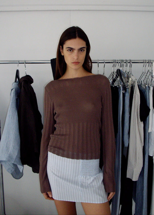 A young woman with long dark hair stands indoors in front of a clothing rack, wearing a brown long-sleeve top and the Shirring Skirt - Mineral Stripe, made from light blue organic cotton. Various Deiji Studios garments hang behind her.