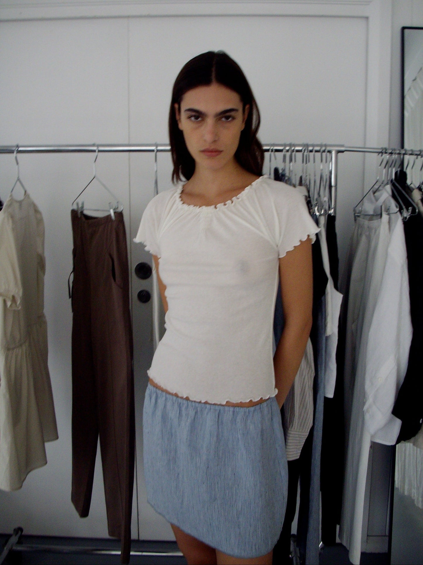 A person with long dark hair stands indoors wearing a white short-sleeve shirt and the Babylock Skirt - Blue Stripe. Behind them, a clothing rack displays french linen garments in neutral tones, suggesting a chic dressing room setting.