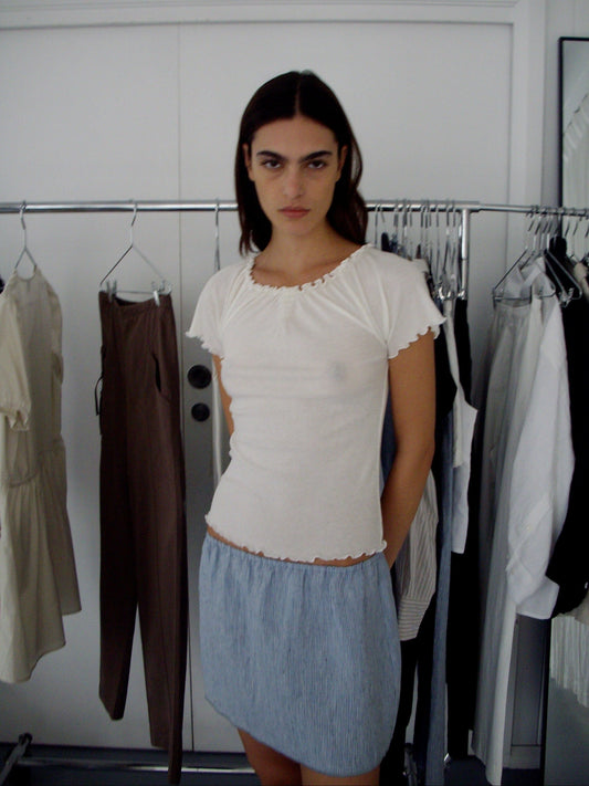 A person with long dark hair stands indoors wearing a white short-sleeve shirt and the Babylock Skirt - Blue Stripe. Behind them, a clothing rack displays french linen garments in neutral tones, suggesting a chic dressing room setting.