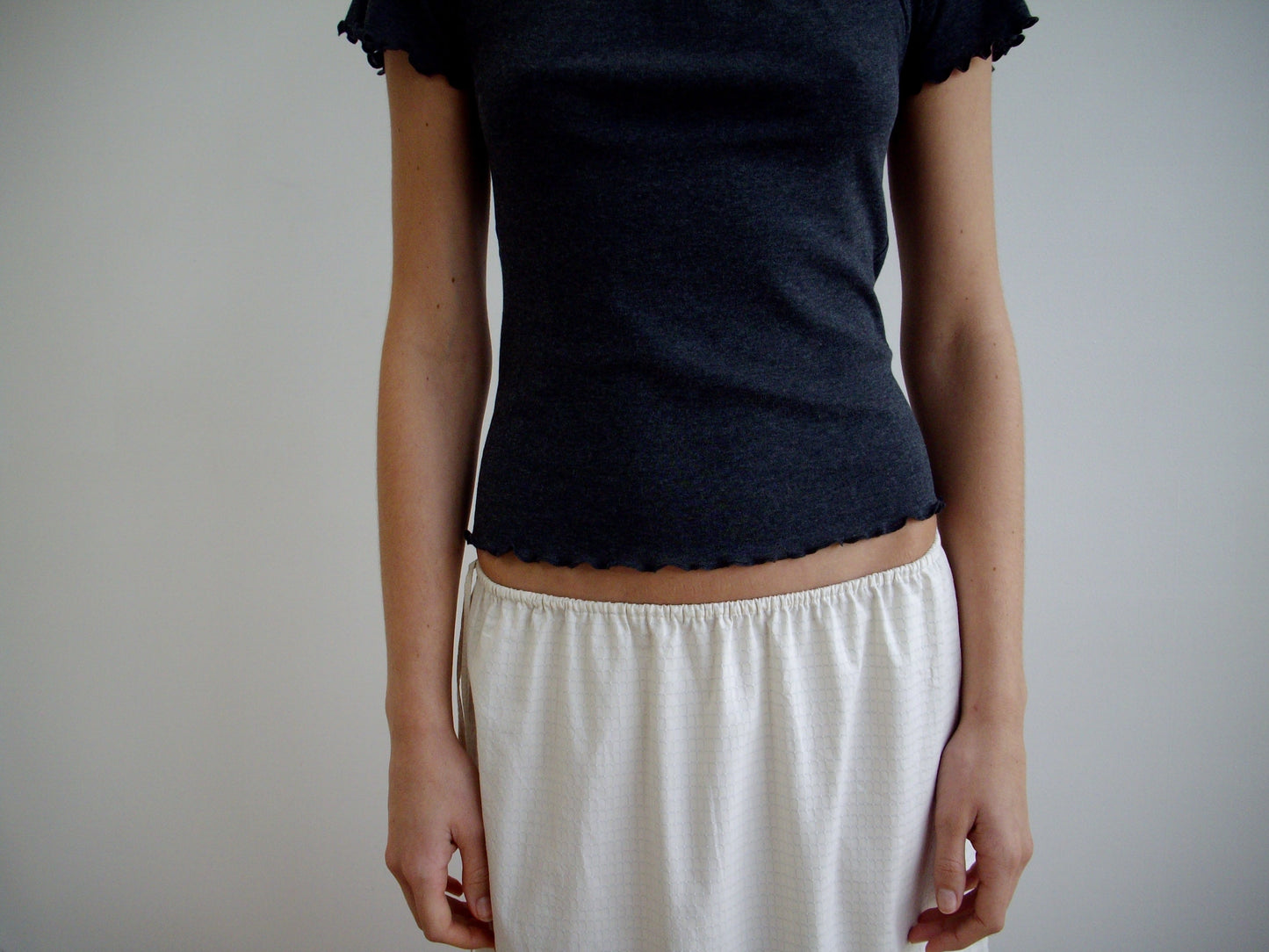 Against a plain background, a person wears the Mid Day Skirt in Bone Check—an organic cotton, loose-fit skirt—paired with a short-sleeved dark gray top. Their arms rest by their sides and their head is out of frame.