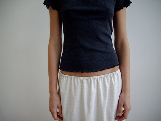 Against a plain background, a person wears the Mid Day Skirt in Bone Check—an organic cotton, loose-fit skirt—paired with a short-sleeved dark gray top. Their arms rest by their sides and their head is out of frame.