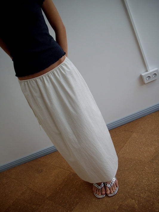 A person stands indoors on a brown floor near a white wall with outlets, wearing a black shirt, the Mid Day Skirt - Bone Check (an organic cotton midi in white), and white sandals.