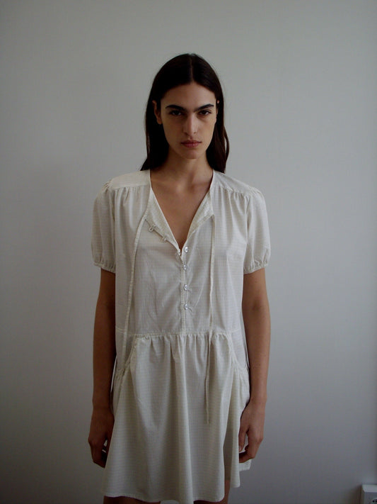 A woman with long dark hair, dressed in the Pocket Mini Dress - Bone Check by Deiji Studios, stands against a plain light wall, gazing at the camera with a neutral expression.