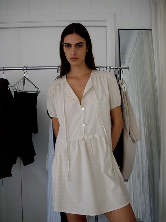 A woman with long dark hair wears the Pocket Mini Dress - Bone Check from Deiji Studios, featuring buttons and pockets, as she stands indoors in front of a clothing rack and a white wall.