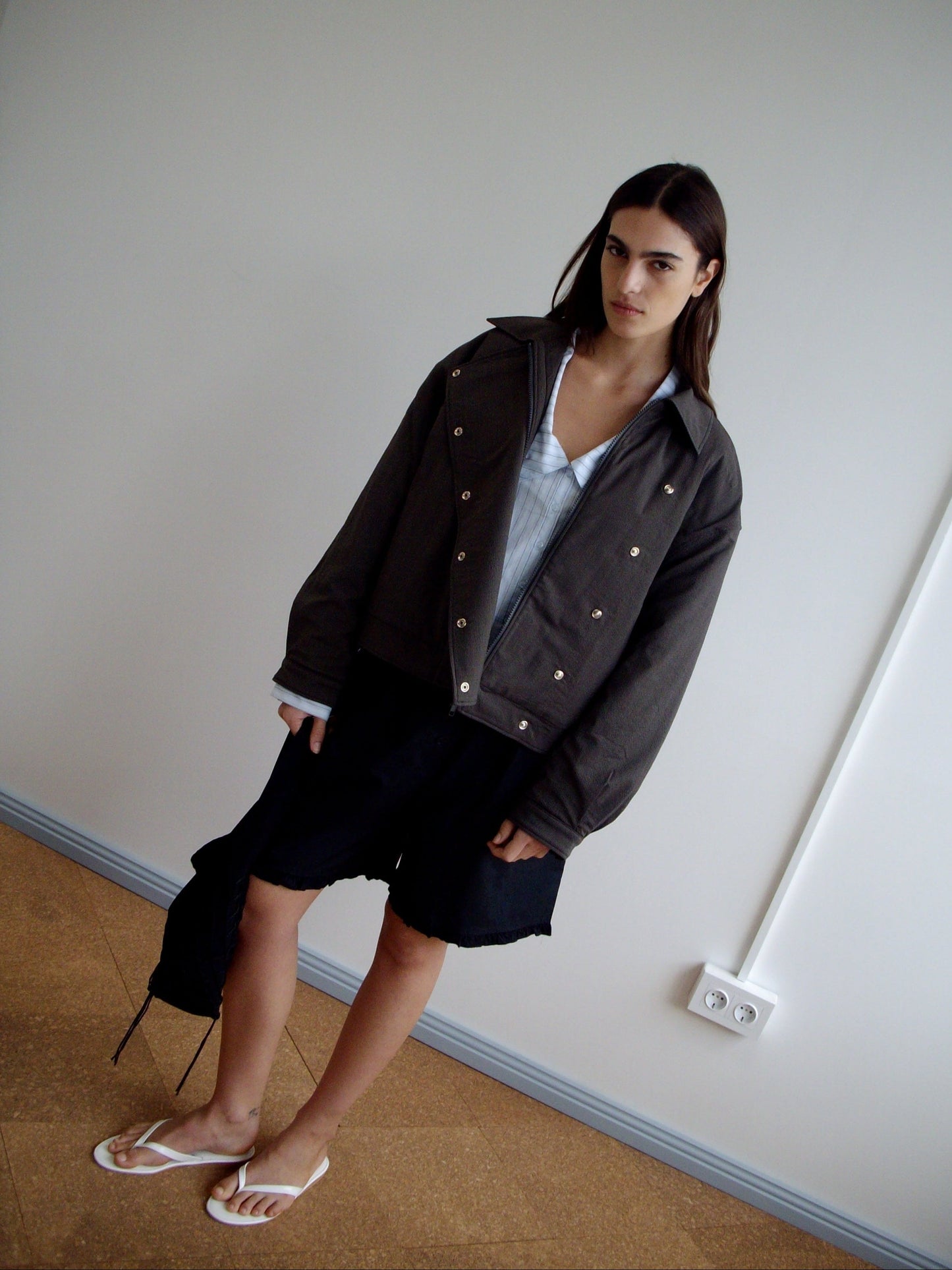 A person with long dark hair stands indoors against a plain wall, wearing a Deiji Studios Lapel Jacket - Brown Stripe over a light blue shirt, black shorts, and white flip-flops, holding a black bag by their side.