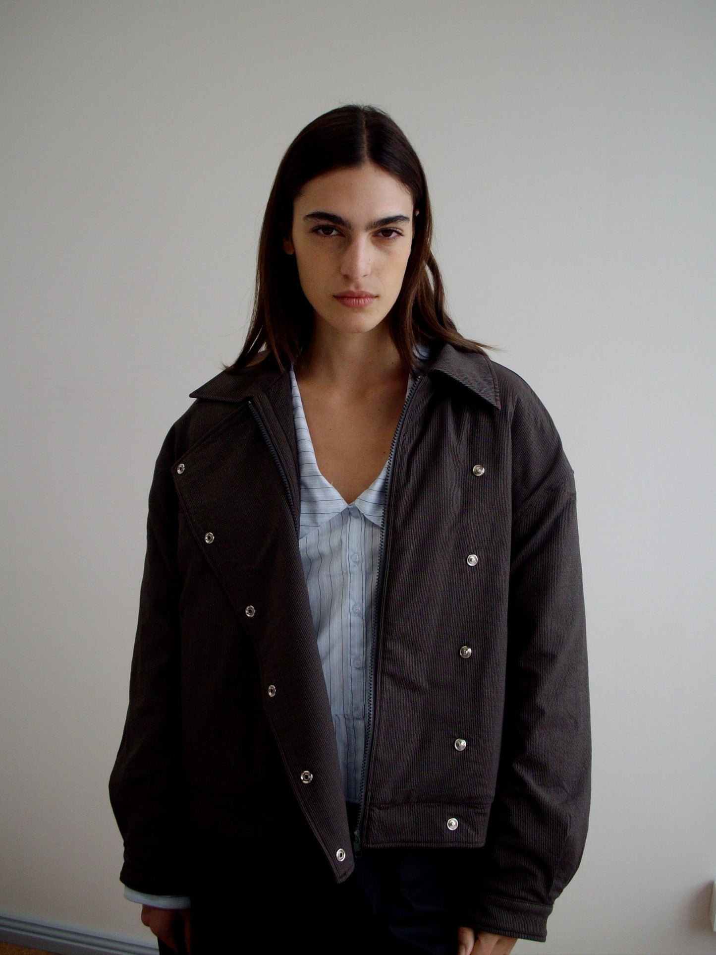 A woman with long brown hair stands indoors against a plain wall, wearing the Lapel Jacket - Brown Stripe over a light blue striped organic cotton shirt. She faces the camera with a neutral expression.