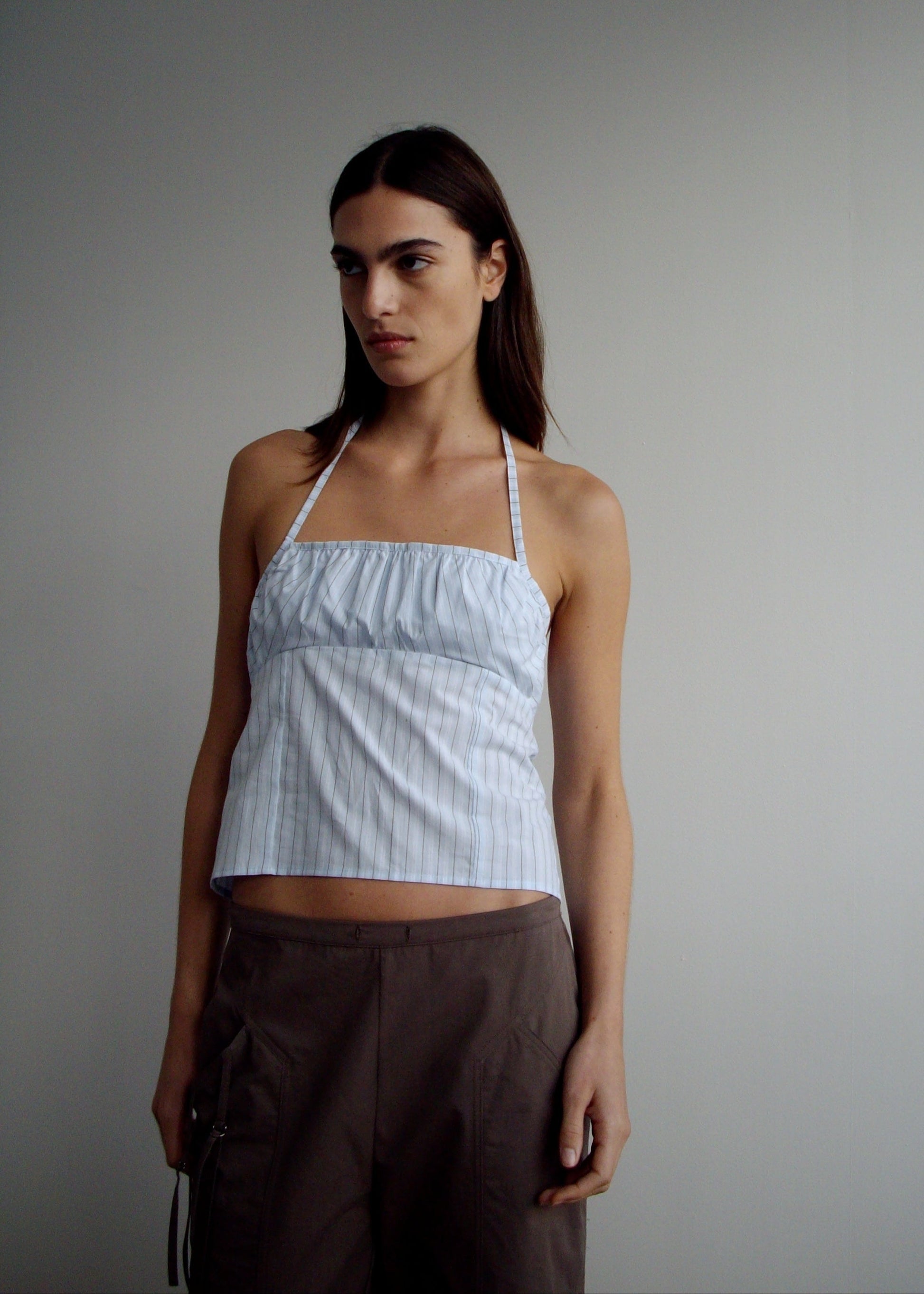 A person with long dark hair is wearing the Strap Top - Mineral Stripe, a light blue pinstriped halter made from organic cotton, paired with brown pants and standing against a plain light gray background.