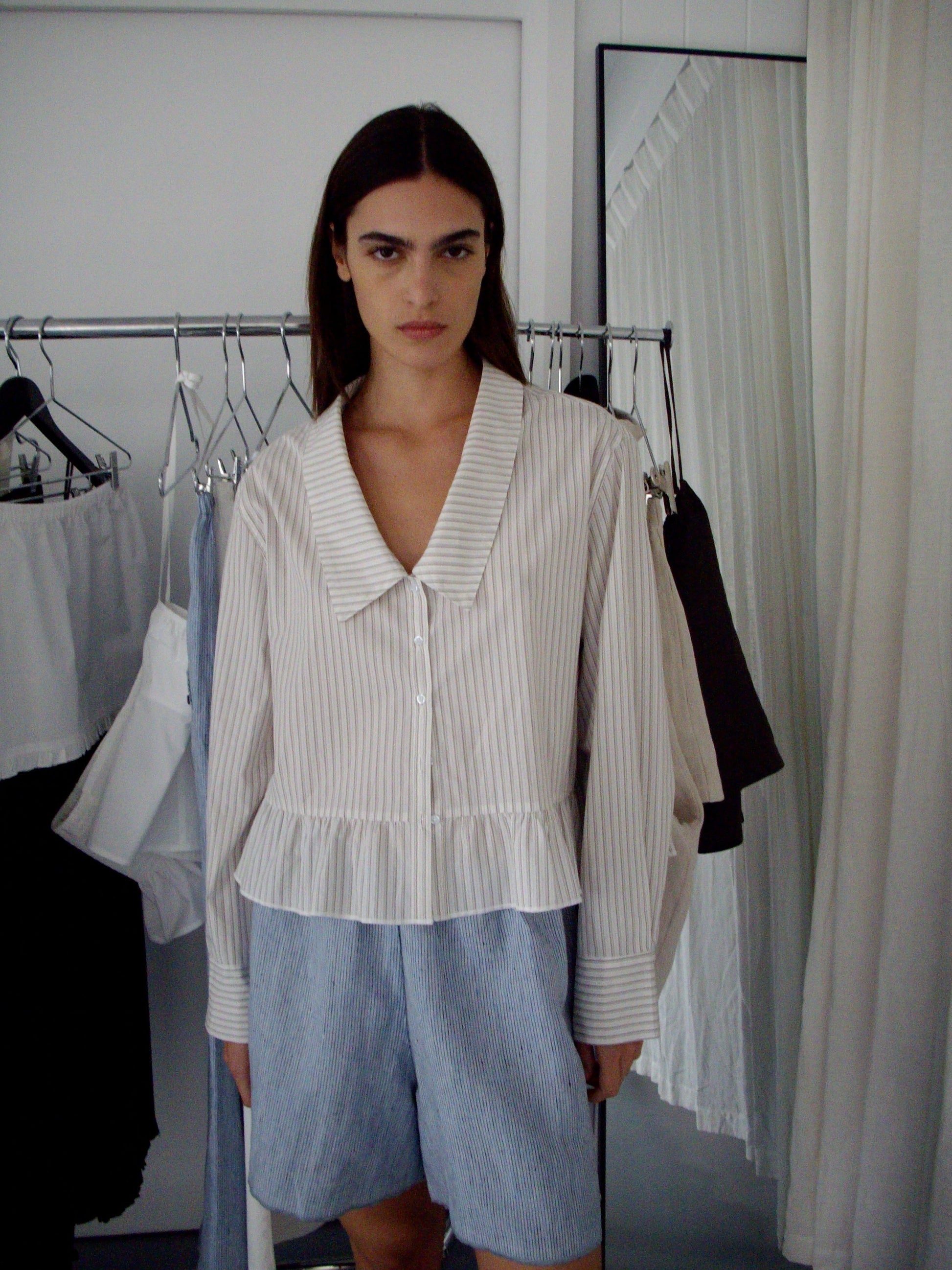 A woman with straight dark hair stands indoors wearing a cream, striped, long-sleeve blouse with a wide collar and ruffled hem, paired with Press Stud Short - Blue Stripe. Behind her are garments on a rack and a mirror.
