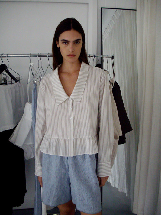 A woman with straight dark hair stands indoors wearing a cream, striped, long-sleeve blouse with a wide collar and ruffled hem, paired with Press Stud Short - Blue Stripe. Behind her are garments on a rack and a mirror.