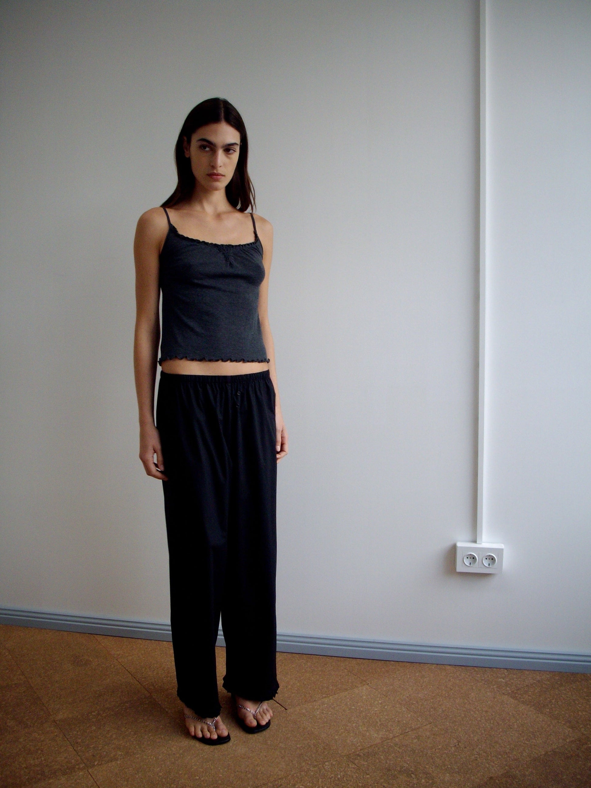 A woman stands indoors against a white wall, wearing the Flounce Tank - Grey by Deiji Studios, loose black pants, and black sandals. She has long, straight hair, a neutral expression, and stands on a brown floor near an electrical outlet.