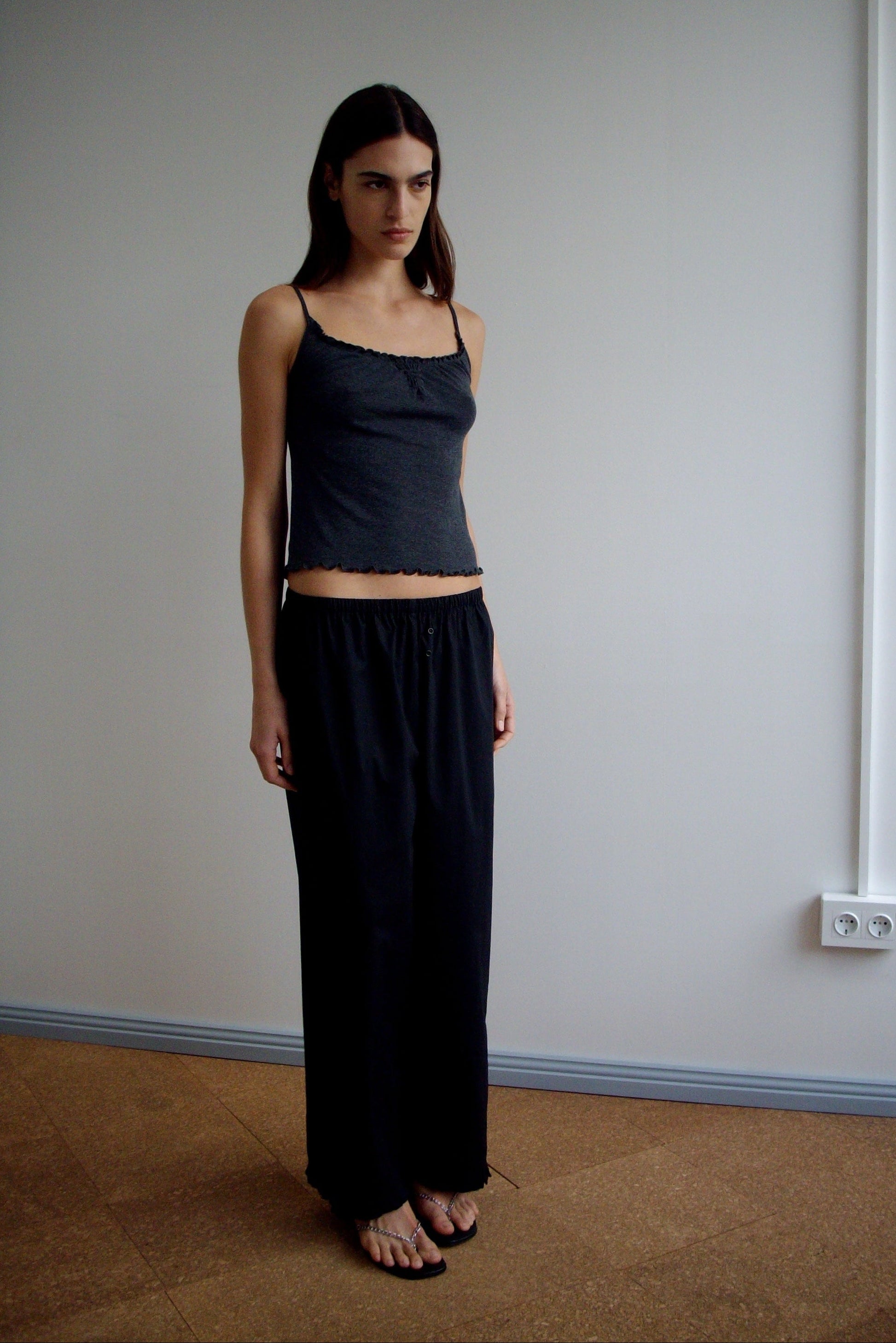 A woman with long dark hair stands indoors against a plain light wall, wearing Deiji Studios’ Flounce Pant - Black and a sleeveless top. She has a neutral expression and her arms rest by her sides in simple elegance.