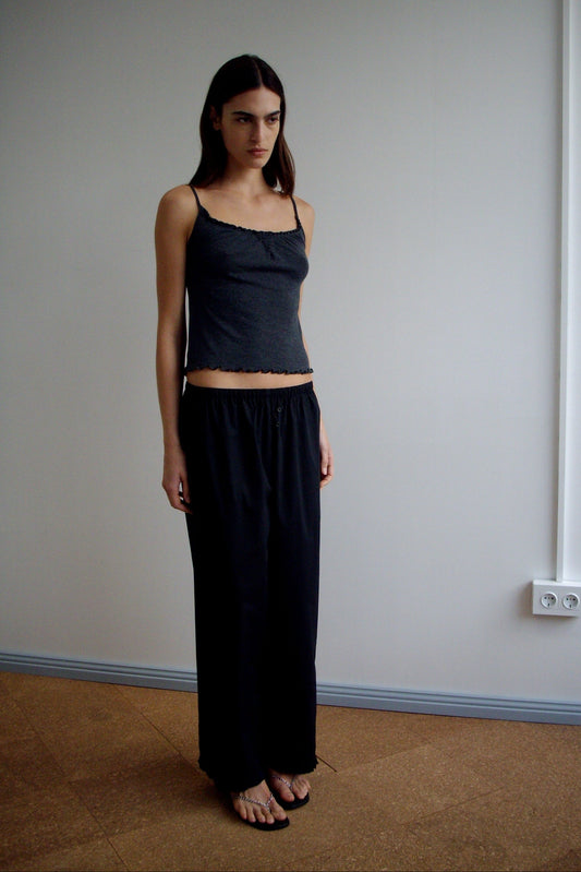 A woman with long dark hair stands indoors against a plain light wall, wearing Deiji Studios’ Flounce Pant - Black and a sleeveless top. She has a neutral expression and her arms rest by her sides in simple elegance.