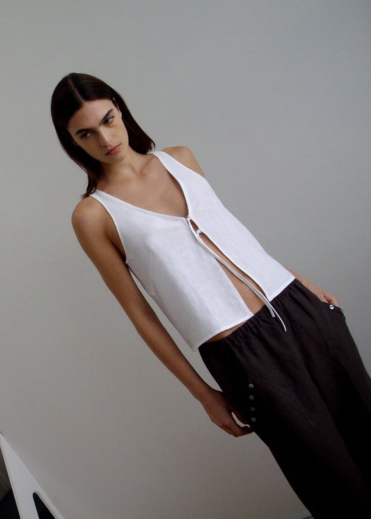 A woman with straight dark hair models the Deiji Studios Two Part Top - White, a sleeveless white linen v-neck with front tie, paired with loose black pants, against a plain light wall, gazing down with a neutral expression.