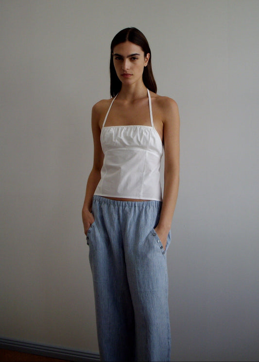 A woman stands against a white wall in the Angle Pocket Pant - Blue Stripe by Deiji Studios, loose and light blue with subtle stripes, paired with a white halter top. Her hands are in her pockets and her straight brown hair frames her neutral expression.