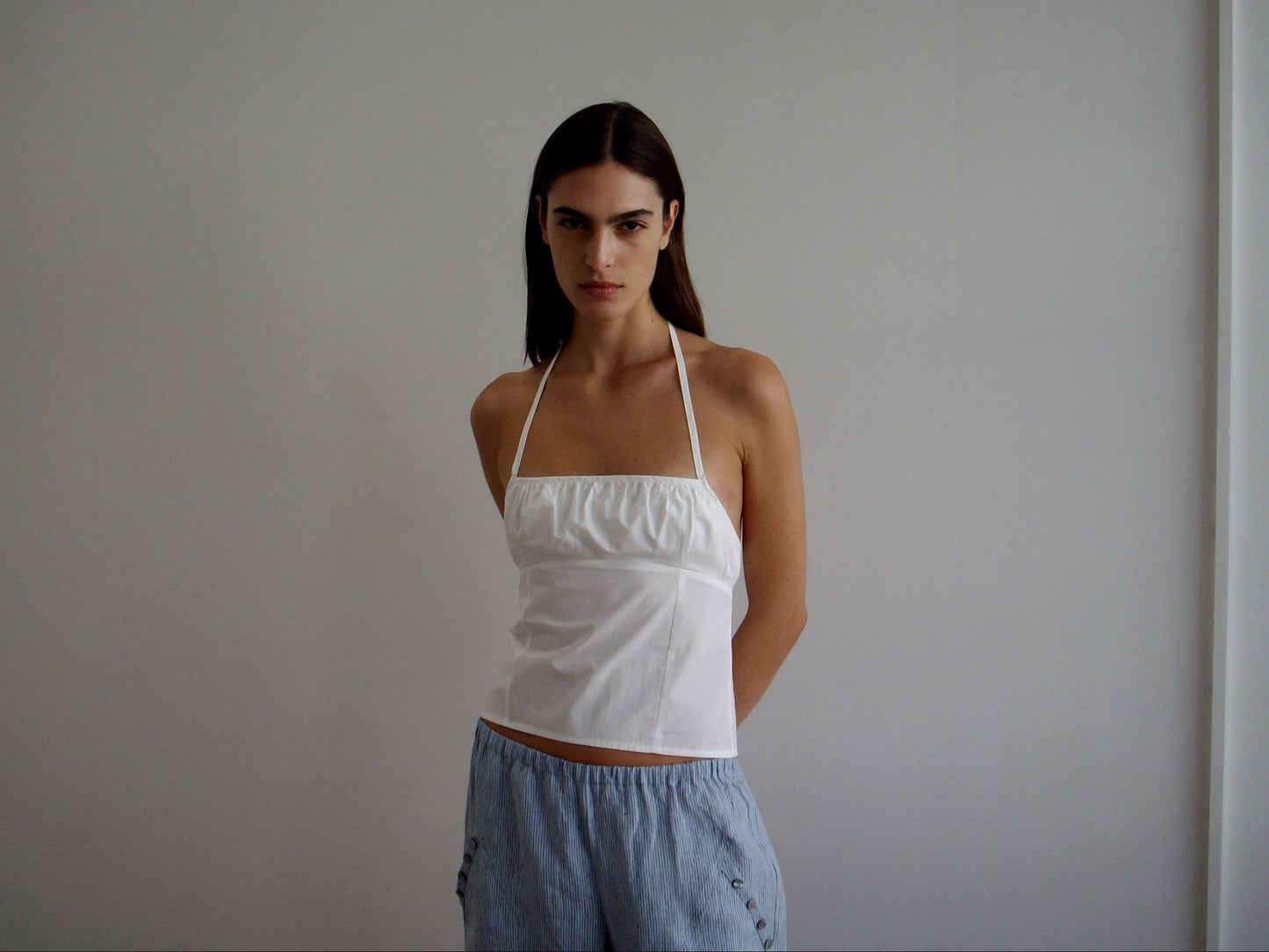 A woman with straight dark hair stands against a plain light wall, wearing the Strap Top - White from Deiji Studios and light blue pants, her arms behind her back and her expression neutral.