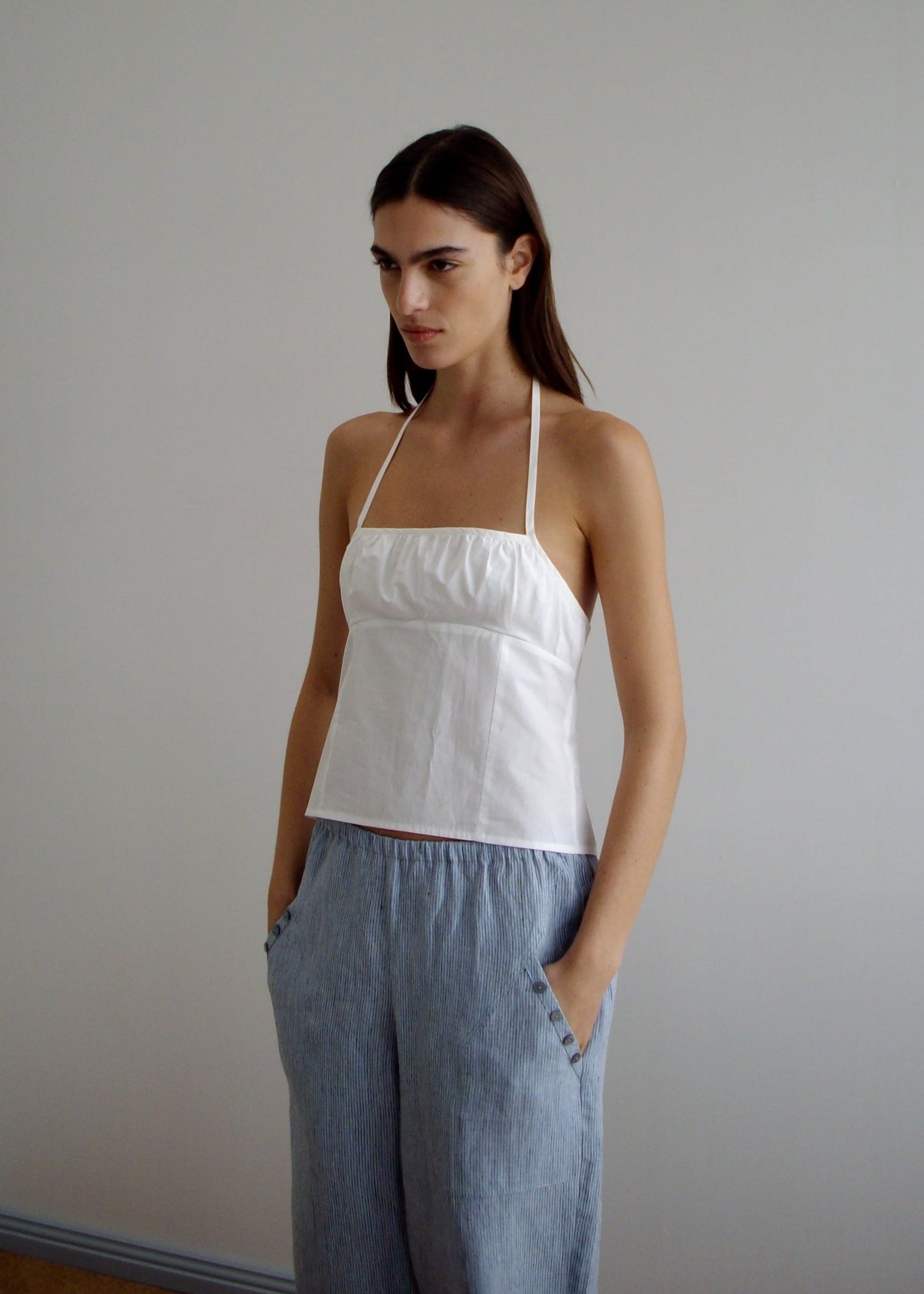 A woman with long brown hair wears the Strap Top - White and loose light blue pants, standing against a plain light-colored wall with her hands in her pockets, looking slightly to the side.