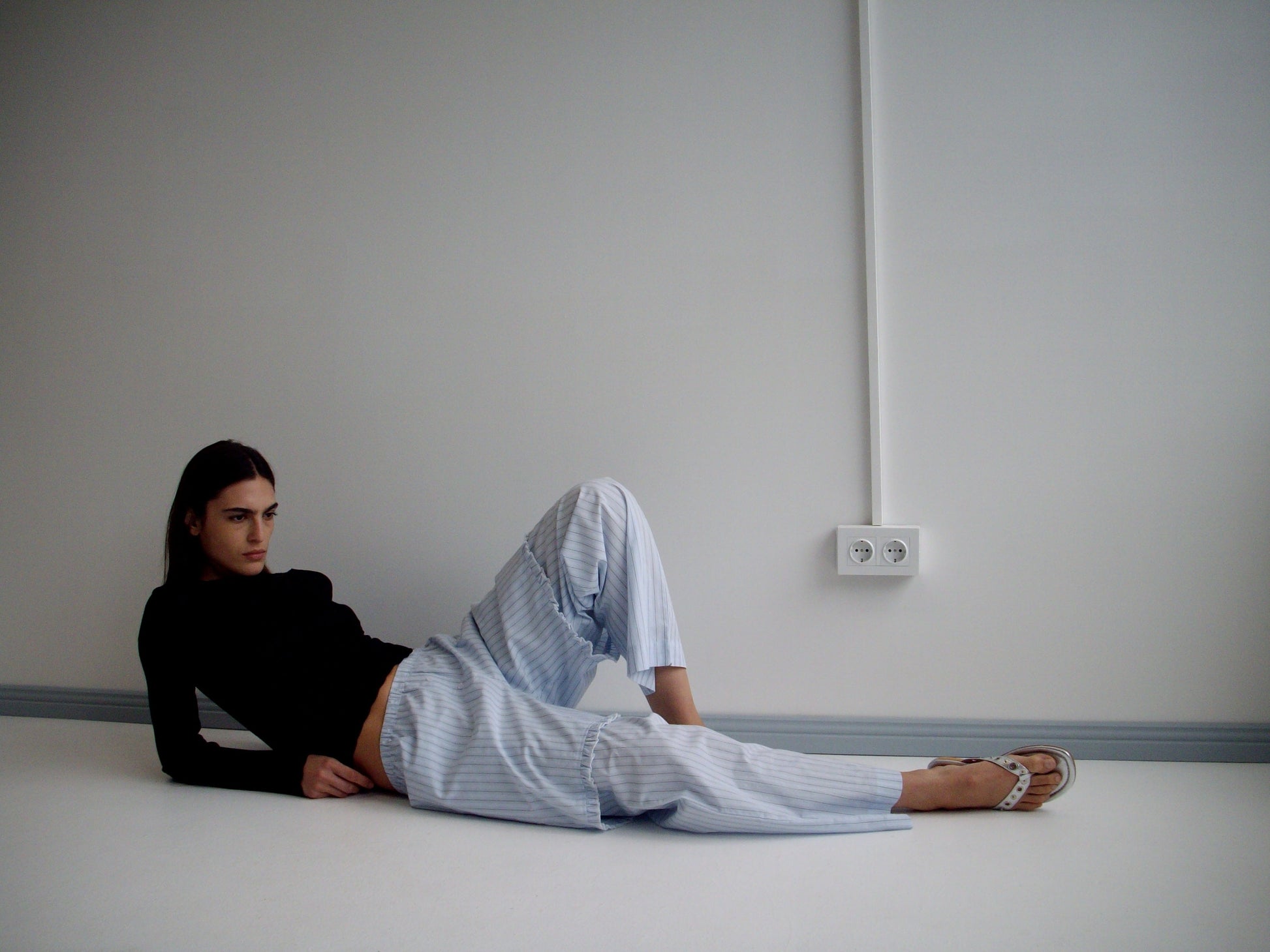 A person relaxes on the floor against a white wall, wearing an organic cotton black long-sleeve top, Deiji Studios’ Ease Trouser in Mineral Stripe, and sandals. An electrical outlet and a visible wire appear on the wall behind them.