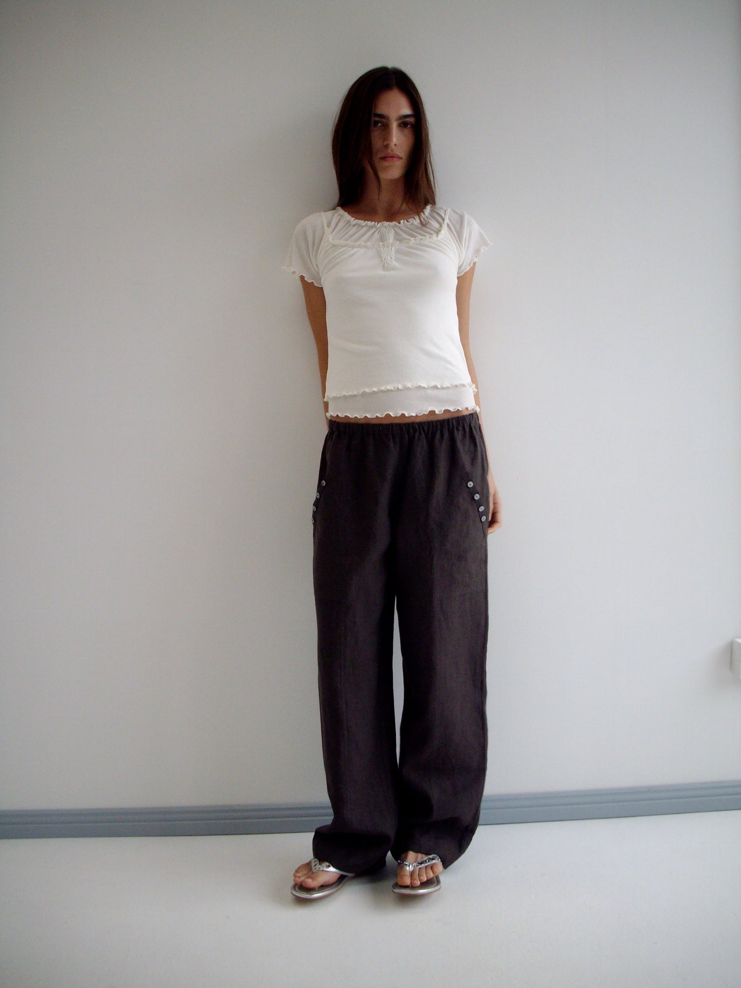 A woman with long dark hair and a neutral expression stands against a white wall wearing the Angle Pocket Pant in Bark with a white short-sleeve top and flat sandals.
