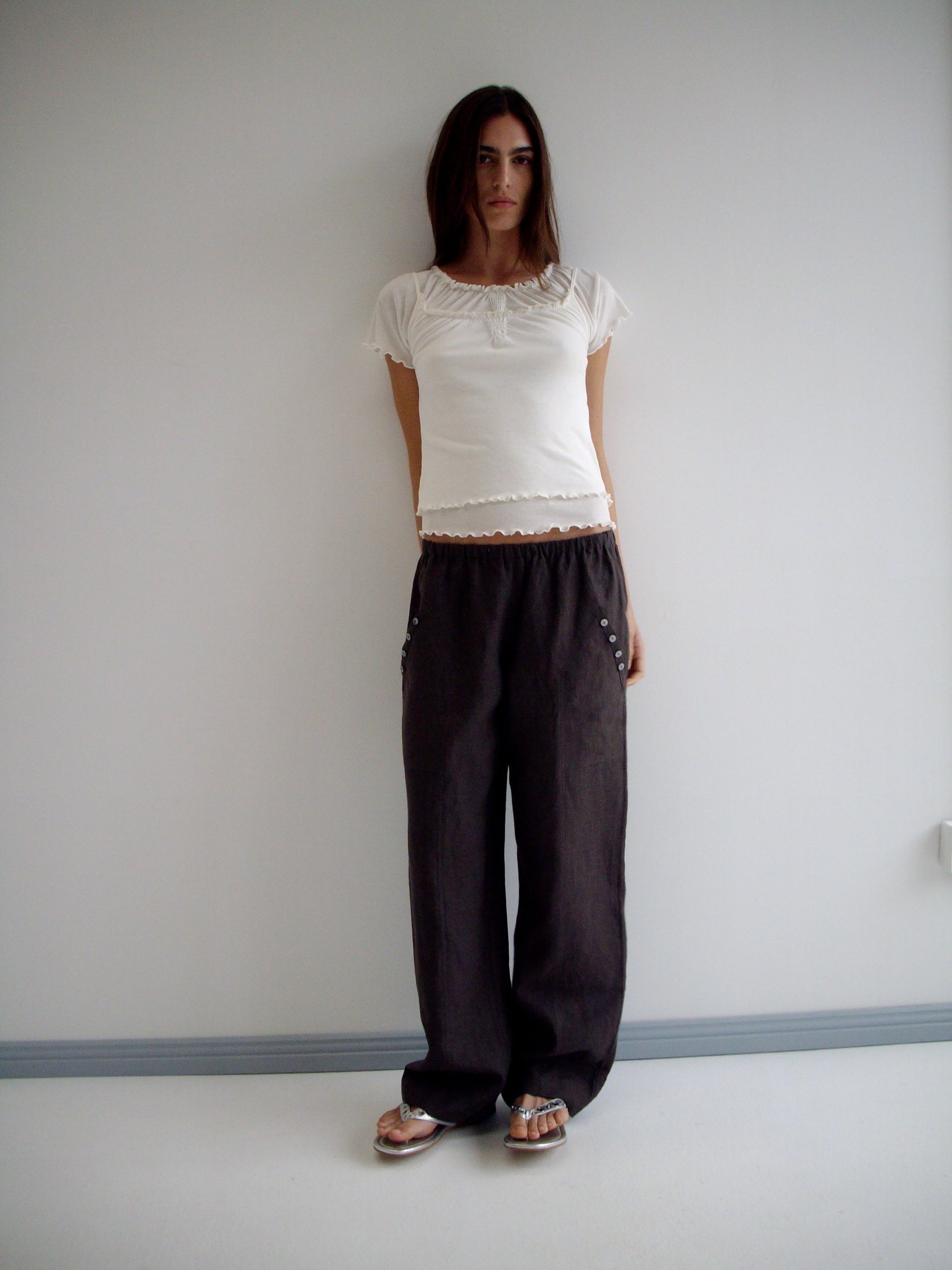 A woman with long dark hair and a neutral expression stands against a white wall wearing the Angle Pocket Pant in Bark with a white short-sleeve top and flat sandals.