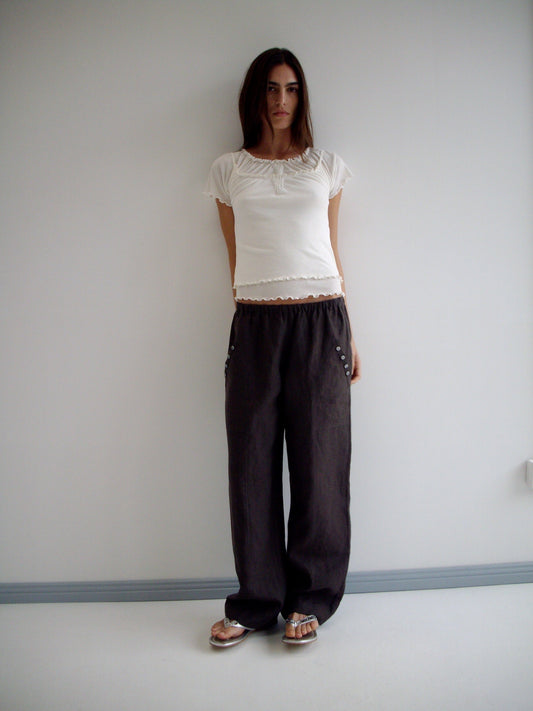 A woman with long dark hair and a neutral expression stands against a white wall wearing the Angle Pocket Pant in Bark with a white short-sleeve top and flat sandals.