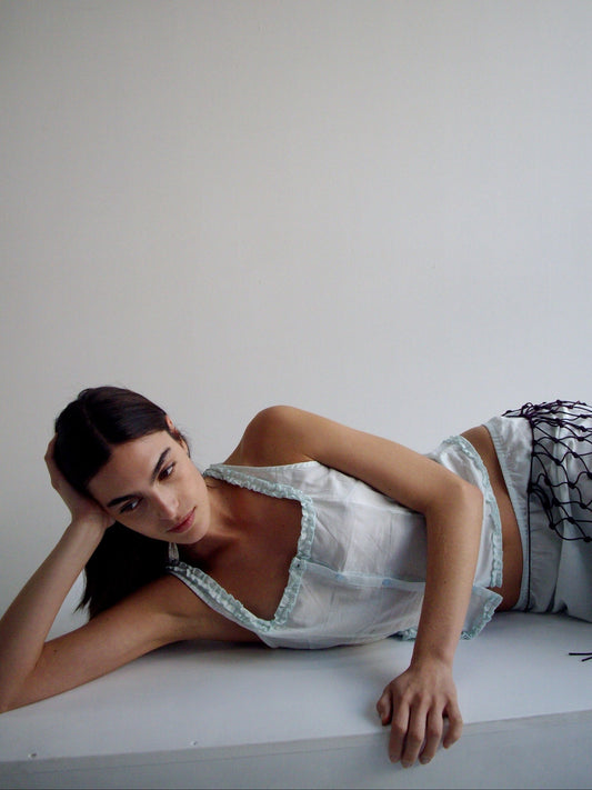 A model reclines on a white surface against a plain background, wearing the Button Frill Top with ruffled details and a white skirt featuring dark cord accents. She rests her head on one hand, gazing softly to the side.