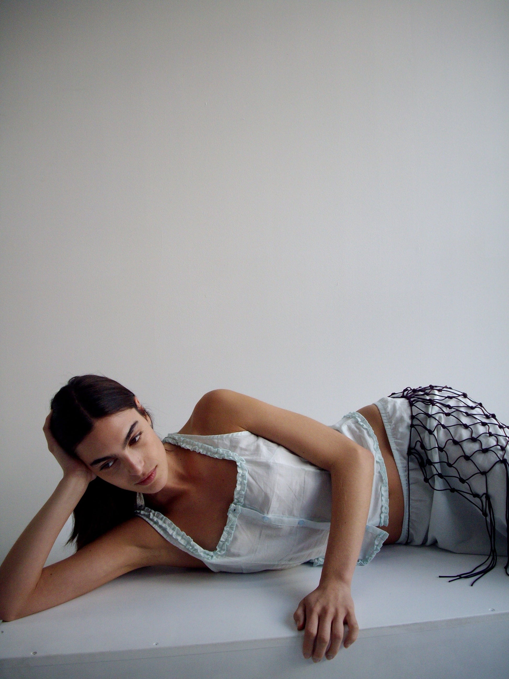 A woman with long dark hair lies on her side against a white background, wearing a light blue halter top, white pants, and a burgundy Macrame Belt draped over one leg, resting her head on her hand and gazing slightly upward.