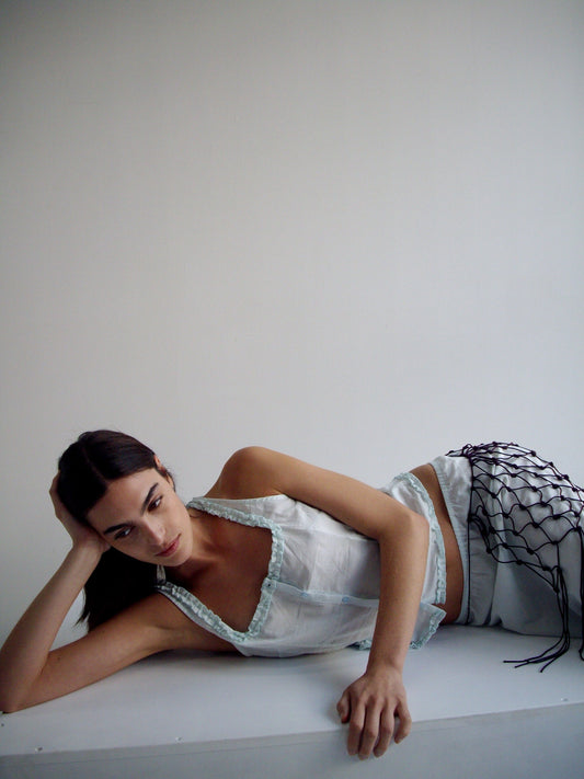 A woman with long dark hair lies on her side against a white background, wearing a light blue halter top, white pants, and a burgundy Macrame Belt draped over one leg, resting her head on her hand and gazing slightly upward.